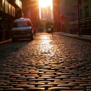May include: A vintage van parked on a cobblestone street in a city. The sun is shining brightly, casting a warm glow on the street and buildings. The van is a light blue color with a white roof and a black bumper. The street is lined with buildings on both sides, and there are trees in the background.