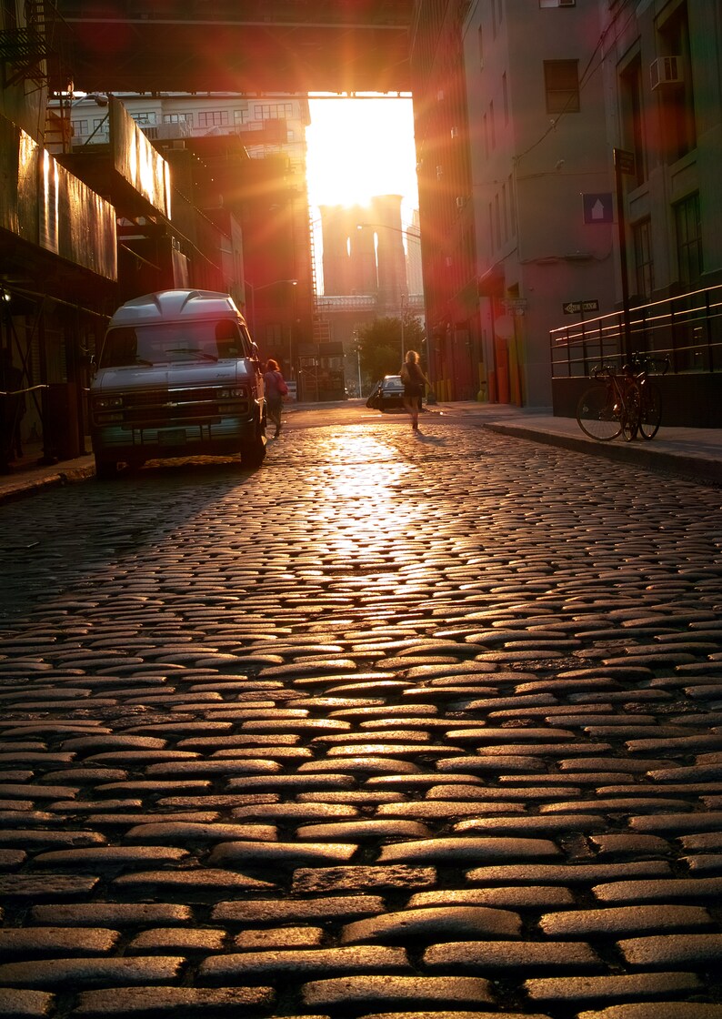 May include: A vintage van parked on a cobblestone street in a city. The sun is shining brightly, casting a warm glow on the street and buildings. The van is a light blue color with a white roof and a black bumper. The street is lined with buildings on both sides, and there are trees in the background.