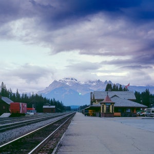 May include: A train station with a red train car on the tracks, a wooden building with a peaked roof, and a mountain range in the background. The sky is cloudy and blue.