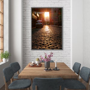 May include: A framed photograph of a cobblestone street in a city. The street is empty except for a car in the distance. The sun is shining brightly and the light is reflecting off the cobblestones.