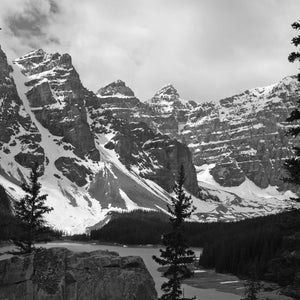 May include: A black and white photograph of a mountain range with snow-capped peaks and a lake in the foreground. The mountains are tall and rugged, and the lake is calm and still. The photo was taken on a cloudy day, and the sky is overcast.