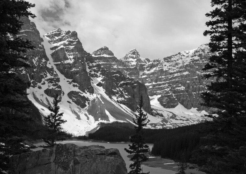 May include: A black and white photograph of a mountain range with snow-capped peaks and a lake in the foreground. The mountains are tall and rugged, and the lake is calm and still. The photo was taken on a cloudy day, and the sky is overcast.
