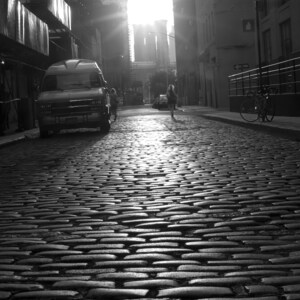 May include: A black and white photo of a cobblestone street in a city. A white van is parked on the left side of the street. A person is walking away from the camera in the middle of the street. The street is lined with buildings on both sides.