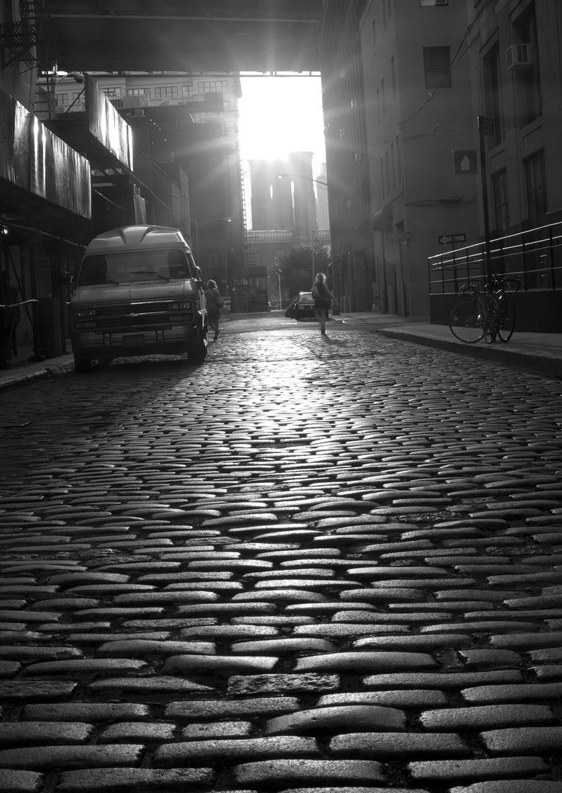 May include: A black and white photo of a cobblestone street in a city. A white van is parked on the left side of the street. A person is walking away from the camera in the middle of the street. The street is lined with buildings on both sides.