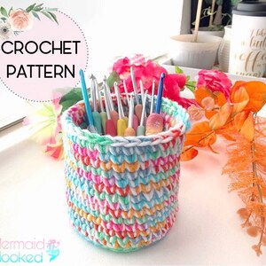 May include: A colorful crochet yarn basket with a white rim, filled with a variety of crochet hooks. The basket is sitting on a white surface with a floral arrangement in the background. The text "CROCHET PATTERN" is visible in the top left corner of the image.