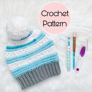 May include: A crocheted hat with a white pom-pom, white, light blue, and grey stripes. The image also shows crochet hooks, a yarn needle, and scissors. The text "Crochet Pattern" is in a pink circle.