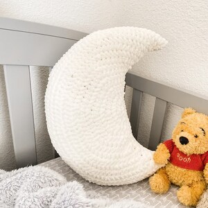 May include: White crocheted crescent moon shaped pillow on a gray crib with a gray and white patterned sheet and a Winnie the Pooh stuffed animal.