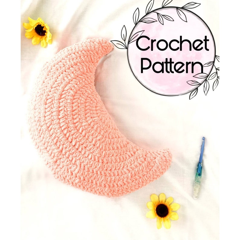 May include: A crochet pattern for a pink crescent moon shaped pillow. The pillow is lying on a white surface with a sunflower and a crochet hook.