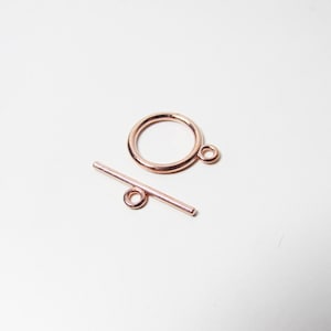 Könnte beinhalten: Roségold-Schließe mit einem runden Ring und einer geraden Stange mit einer Schlaufe an einem Ende.