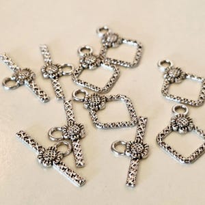 Può includere: Una collezione di chiusure a T in metallo color argento per la creazione di gioielli. Ogni chiusura è composta da una barra con motivo a tratteggio incrociato e un connettore a forma di girasole, e una cornice quadrata con un dettaglio floreale.