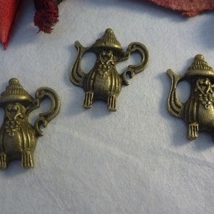 Puede incluir: Tres encantos de tetera de color dorado antiguo con detalles ornamentados. Cada encanto presenta una tetera con tapa y asa, y un diseño decorativo en el cuerpo.
