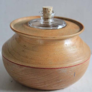 May include: A wooden jar with a lid and a glass bottle inside. The jar is light brown with a red stripe around the middle. The lid is also made of wood and has a cork stopper on top of the glass bottle.