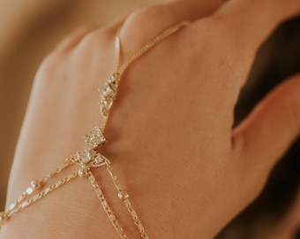 Puede incluir: Un brazalete de cadena dorada con un diseño delicado y pequeñas piedras transparentes. El brazalete se lleva en la muñeca y tiene un bucle para el dedo.