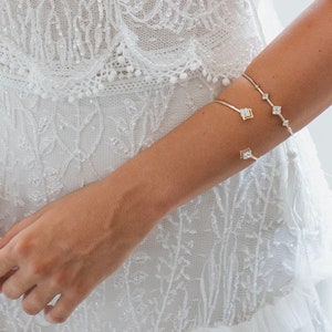 May include: A gold-toned bracelet with square and round clear gemstones is worn on an arm. The arm is adorned with a second gold bracelet. The person is wearing a white, embellished dress with a detailed bodice and a tiered skirt.