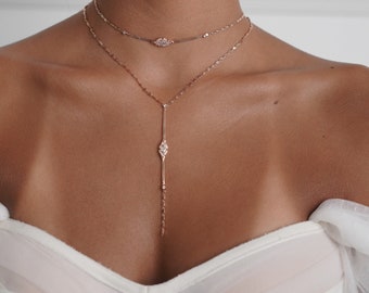 May include: A woman wearing a white off-the-shoulder top with a delicate gold chain necklace and a longer pendant necklace with a diamond-shaped pendant.