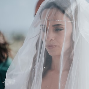 May include: A bride wearing a white veil with a delicate beaded headband. The veil is sheer and covers her face, creating a soft, ethereal look.