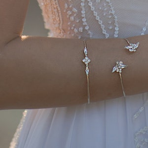 May include: A bride's arm adorned with delicate silver bracelets and a ring. The bracelets feature floral and geometric designs. The bride is wearing a white, beaded wedding dress with a V-neckline.