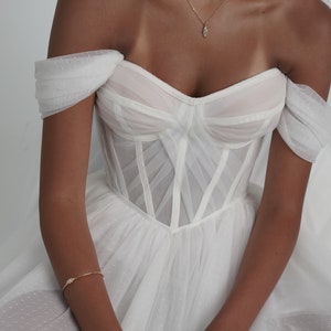 May include: A white off-the-shoulder wedding dress with a fitted bodice and a flowing skirt. The bodice features a corset-style design with delicate straps and a sweetheart neckline. The skirt is made of tulle and has a dotted pattern.