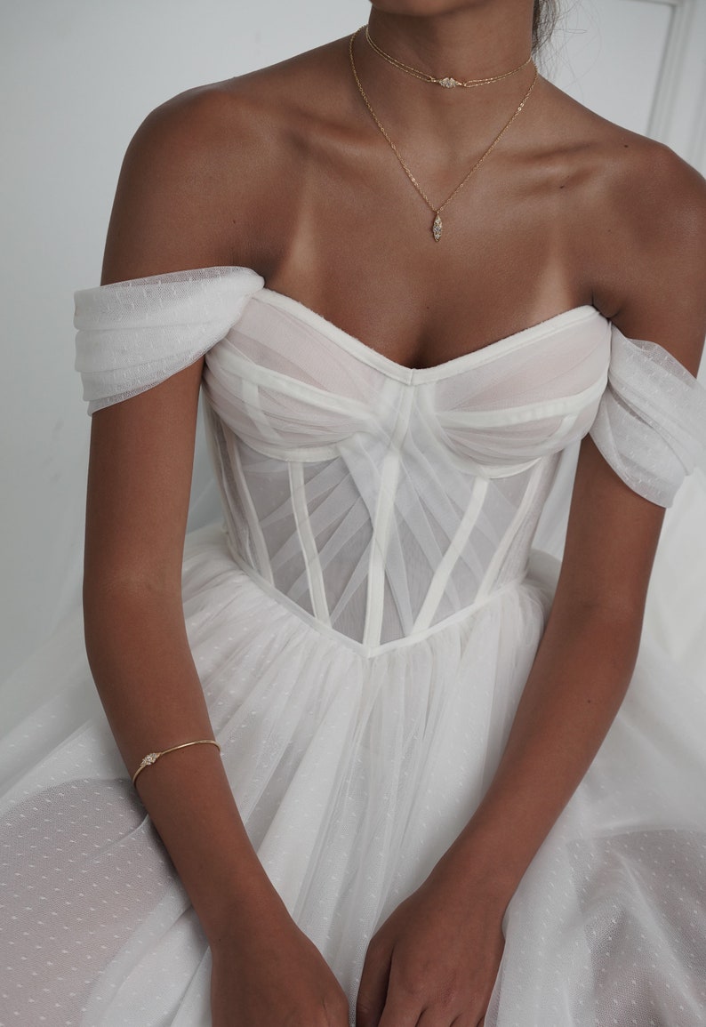 May include: A white off-the-shoulder wedding dress with a fitted bodice and a flowing skirt. The bodice features a corset-style design with delicate straps and a sweetheart neckline. The skirt is made of tulle and has a dotted pattern.