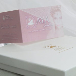 May include: A white box with a gold logo that says "AYA Jewellery" and the website "AYAJewellery.etsy.com". The box is sitting on a white surface. A pink card with the same logo and website is sitting on top of the box.