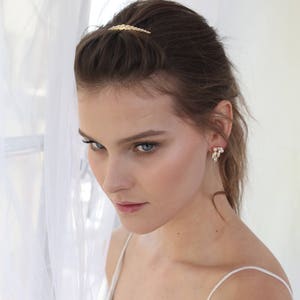 May include: A woman wearing a white bridal gown with a gold hair clip and pearl earrings. The gown has a lace bodice and thin straps.