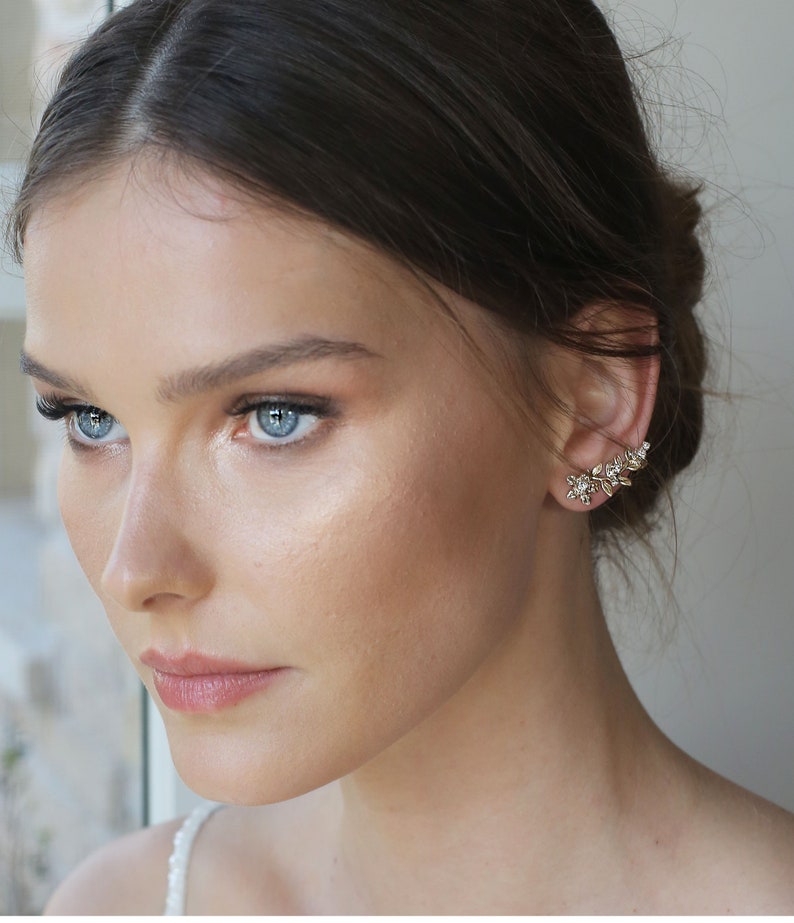 May include: A woman with long brown hair wearing a pair of gold ear climbers with small white stones. The ear climbers are shaped like a vine with leaves and flowers.