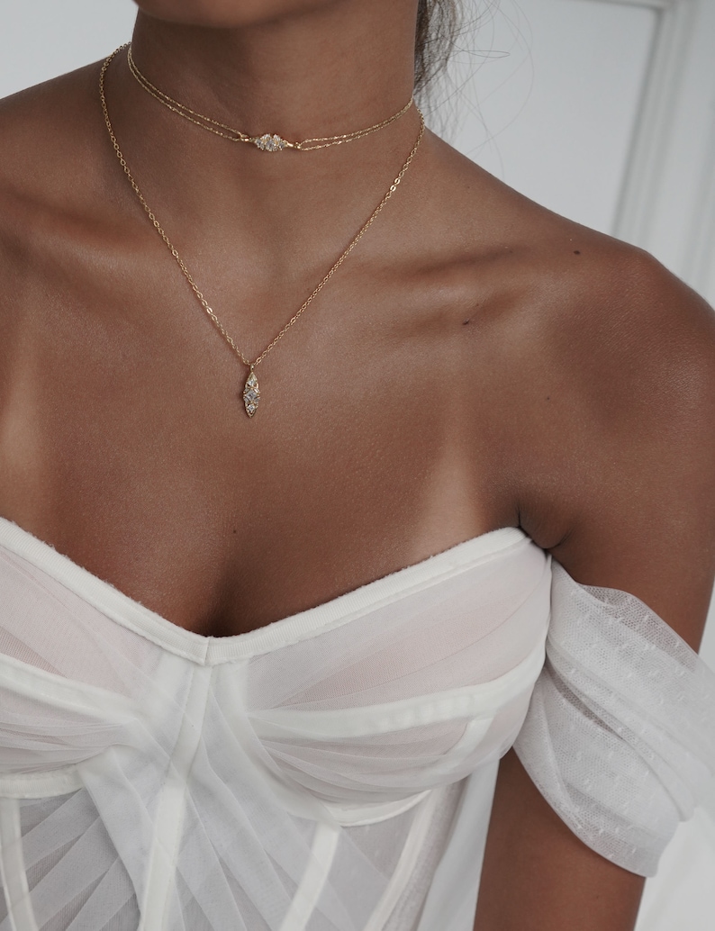 May include: A woman wearing a white off-the-shoulder top with a gold chain necklace and a gold choker necklace with a small diamond-shaped pendant.