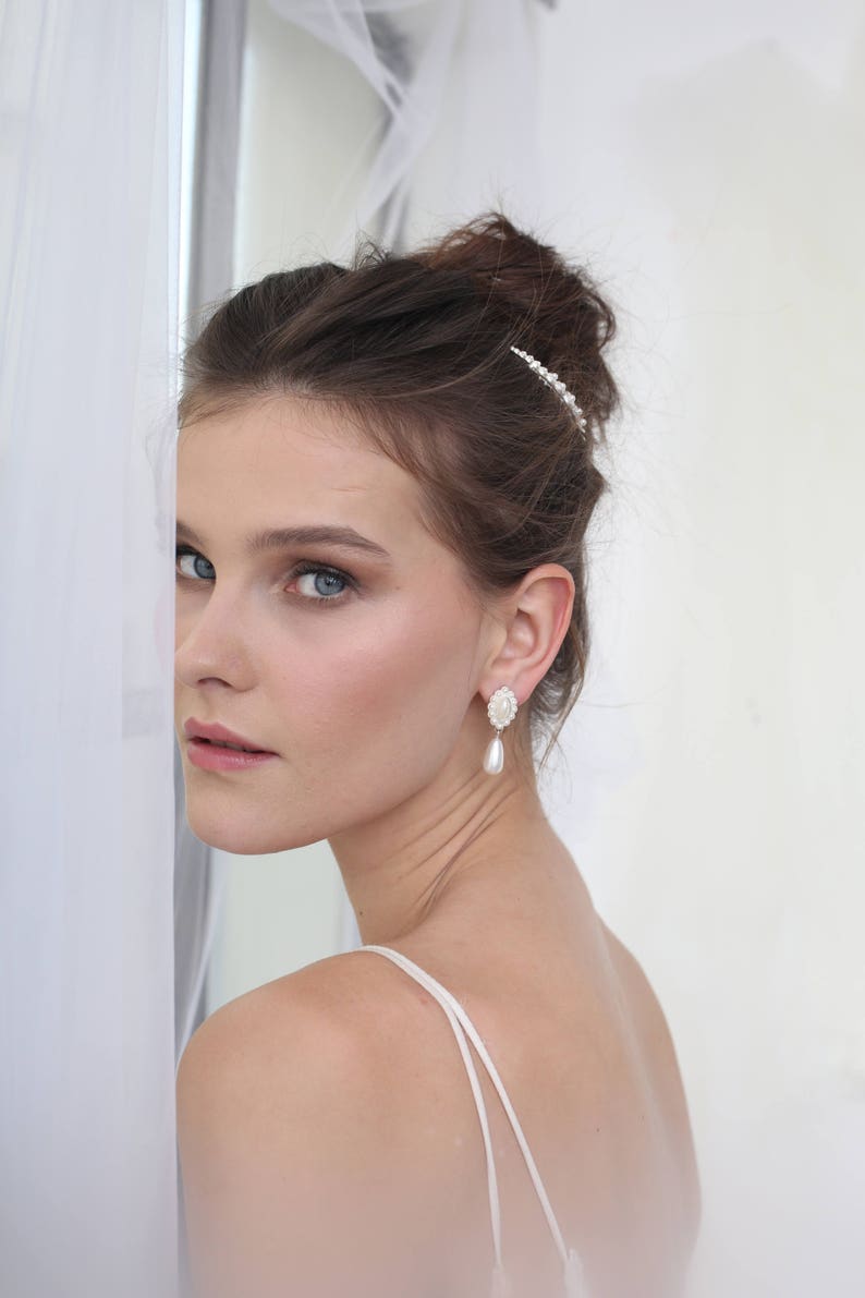 May include: A woman with brown hair wearing a white dress and pearl earrings. She is looking off to the side and has a pearl hair comb in her hair.