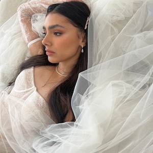 May include: A woman wearing a white lace top and a veil, with a delicate necklace and earrings. She has long dark hair and is lying down.