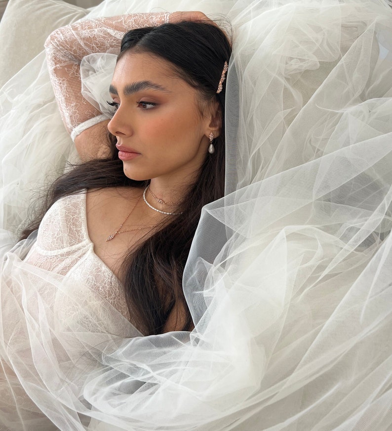 May include: A woman wearing a white lace top and a veil, with a delicate necklace and earrings. She has long dark hair and is lying down.