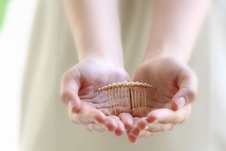 May include: A gold hair comb with pearl accents. The comb is held in the palms of two hands.