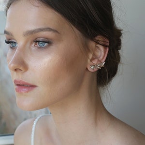 May include: A woman with long brown hair wearing a white top and gold ear cuff earrings with small crystals.
