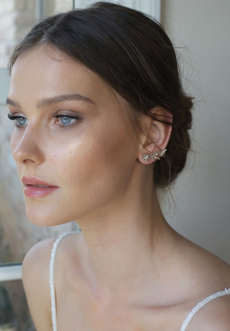 May include: A woman with long brown hair wearing a white top and gold ear cuff earrings with small crystals.
