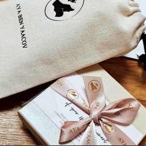 Op de afbeelding: Een geschenkdoos met een roze lint en de tekst "AYA BEN YAACOV". Een beige trekkoordtas staat op de achtergrond. Het lint heeft gouden accenten en de woorden "of your moment".