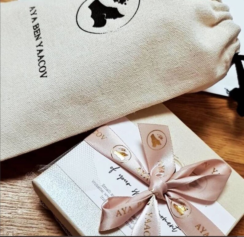 Op de afbeelding: Een geschenkdoos met een roze lint en de tekst "AYA BEN YAACOV". Een beige trekkoordtas staat op de achtergrond. Het lint heeft gouden accenten en de woorden "of your moment".