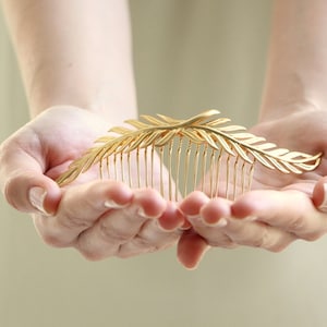 May include: A gold leaf-shaped hair comb with a delicate design. The comb is held in the palms of two hands.