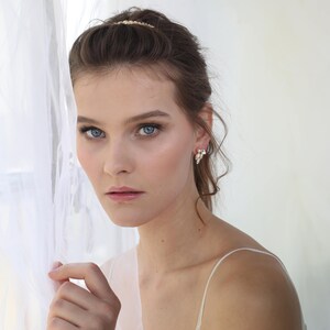 May include: A woman with long brown hair wearing a white top and pearl earrings. She is standing near a white sheer curtain.