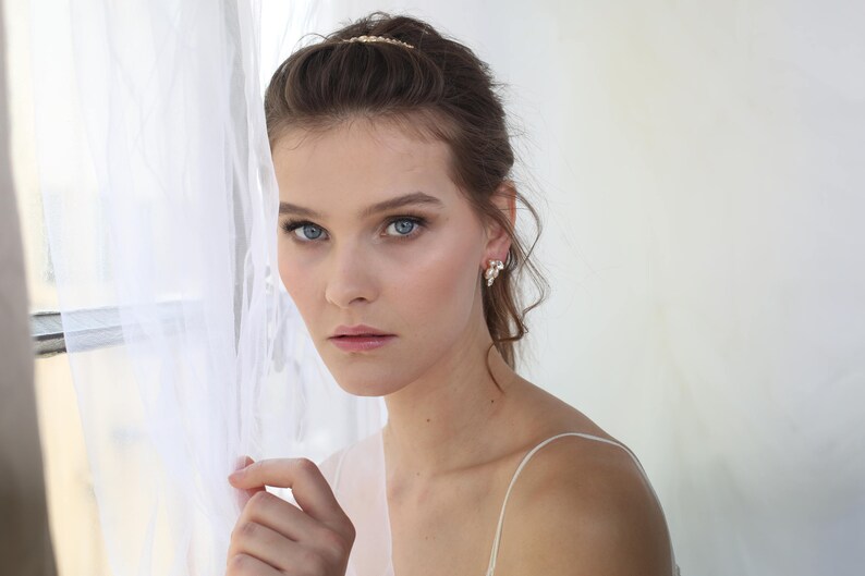 May include: A woman with long brown hair wearing a white top and pearl earrings. She is standing near a white sheer curtain.