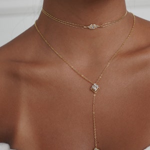 May include: A woman wearing a white top with a gold chain necklace featuring two pendants with clear stones. The necklace is layered with a delicate gold chain with a small diamond-shaped pendant.