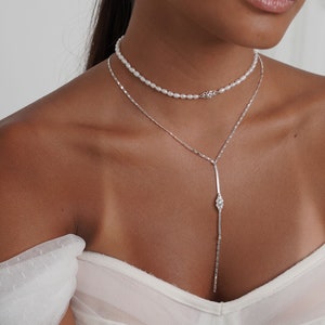 Op de afbeelding: Elegante sieradenset met een parel choker ketting en een lange zilveren hanger ketting met een diamant-achtig accent. Bijpassende parel oorbellen maken de look compleet. Het model draagt een witte jurk.