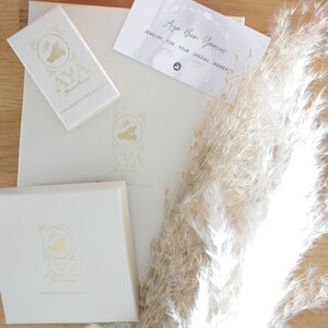 May include: A close-up of two small, clear crystal earrings with gold settings. The earrings are on a wooden surface with a white box and a business card that reads "Aya Ben Yacov Jewelry for your special moments."