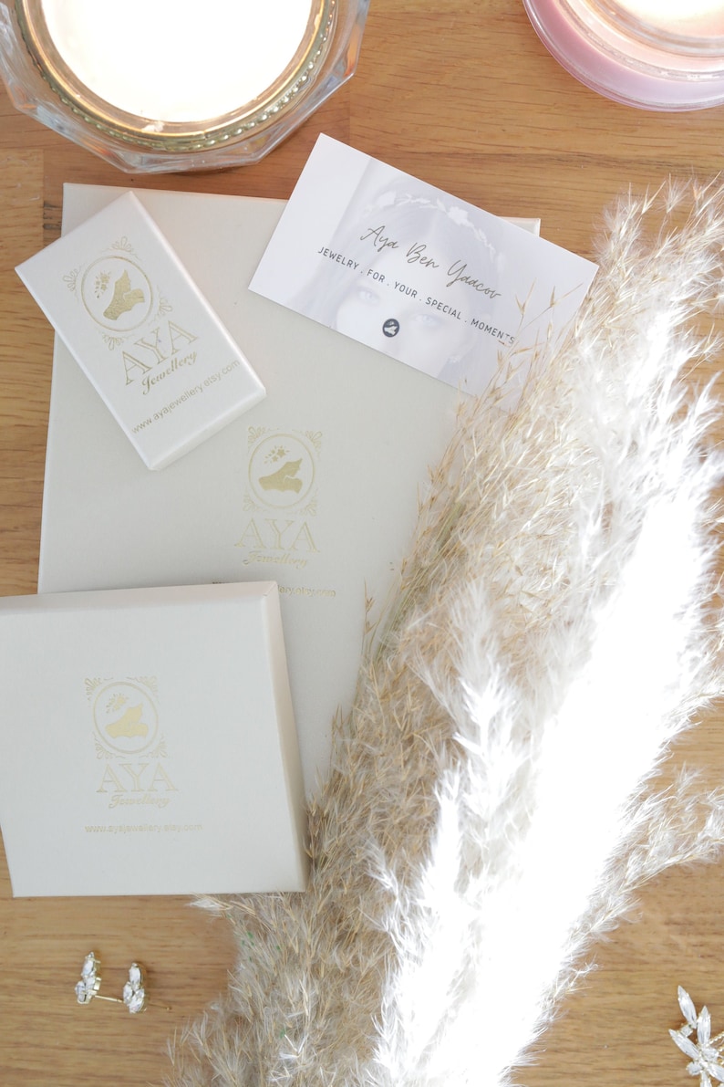 May include: A close-up of two small, clear crystal earrings with gold settings. The earrings are on a wooden surface with a white box and a business card that reads "Aya Ben Yacov Jewelry for your special moments."