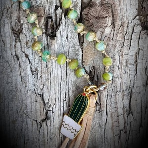 May include: A green and gold beaded necklace with a cactus charm and a brown tassel. The necklace is on a rustic wooden background.