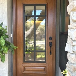 May include: A wooden front door with a glass window pane featuring a black grid design. The door has a dark handle and lockset. A welcome mat sits at the base of the door. The door is framed by a light-colored stone wall and a concrete planter with greenery.