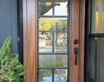 May include: A wooden front door with a black metal grid window pane. The door has a black door handle and a black door lock. The door is surrounded by a black frame.