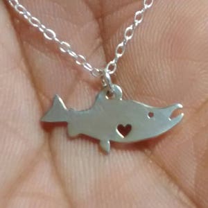 May include: A silver necklace with a delicate fish pendant featuring a small heart cutout. The pendant is on a silver chain.