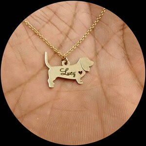 May include: Gold necklace with a dachshund pendant. The pendant has the name "Lucy" engraved on it and a small heart cutout.