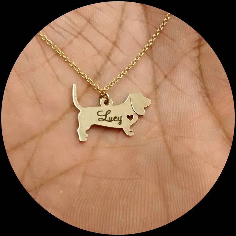 May include: Gold necklace with a dachshund pendant. The pendant has the name "Lucy" engraved on it and a small heart cutout.