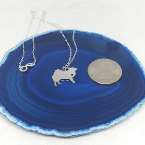May include: A silver necklace with a small, detailed silhouette of a Pomeranian dog charm. The charm is about the size of a US quarter. The necklace is displayed on a blue agate slice.