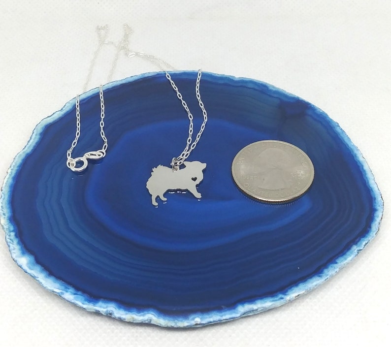May include: A silver necklace with a small, detailed silhouette of a Pomeranian dog charm. The charm is about the size of a US quarter. The necklace is displayed on a blue agate slice.